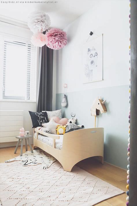 اتاق کودک مدرن با دیوارهای سفید و خاکستری و تخت چوبی که سقف آن با پام پام های کاغذی رنگارنگ تزیین شده است